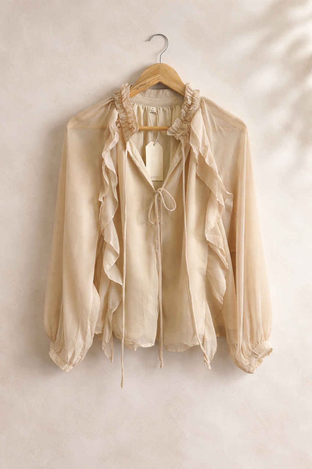 Beige blouse with ruffled details on a wooden hanger against a beige wall.